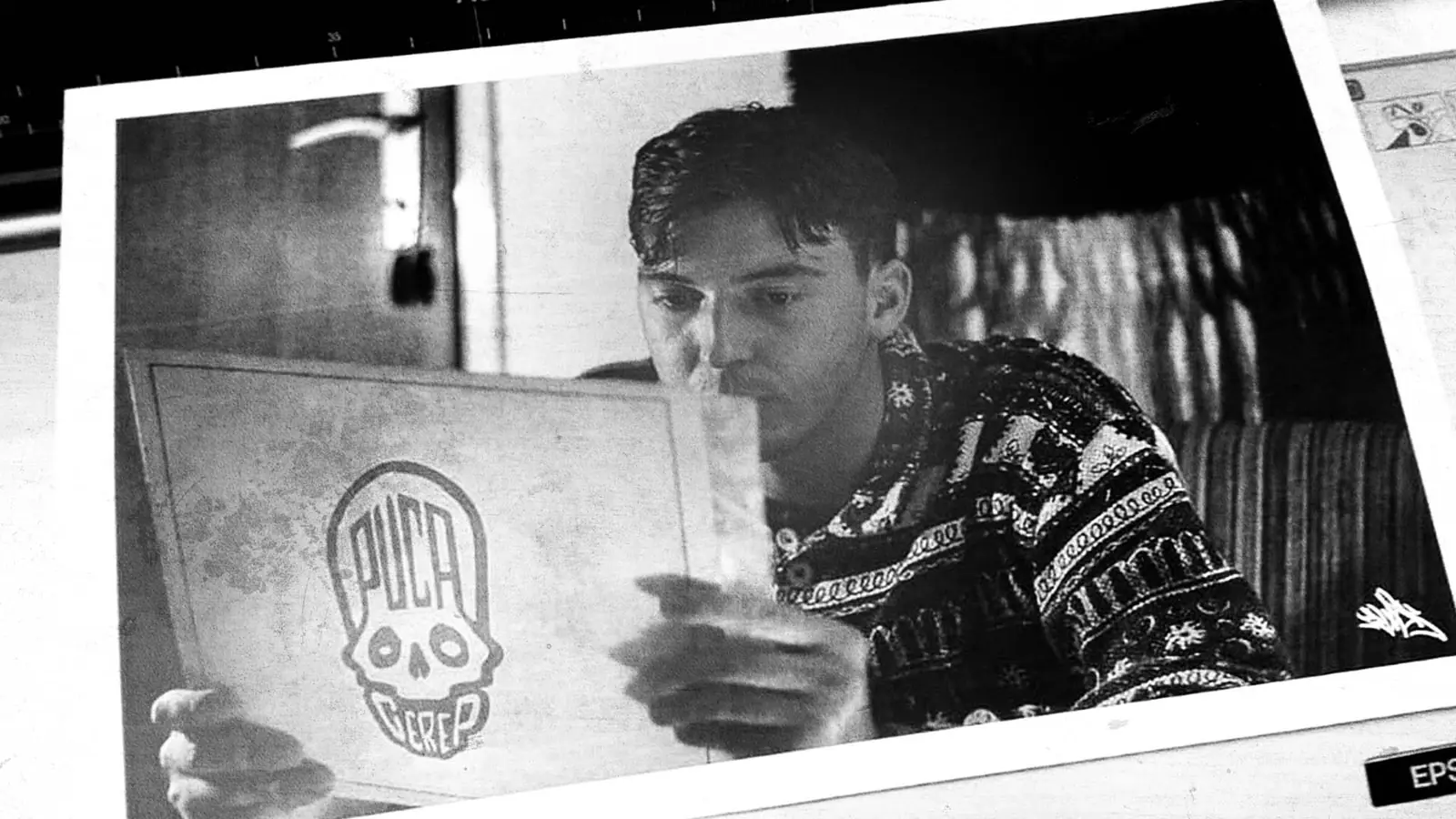 Black and white print photo of a young man holding a board with the Puca Cerep skull logo